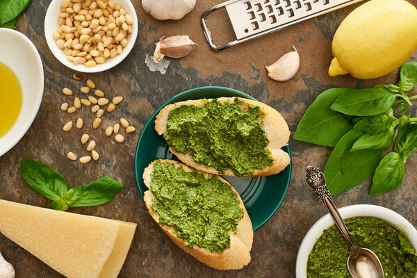 top view of baguette slices with pesto sauce on plate near fresh ingredients and cooking utensils on stone surface