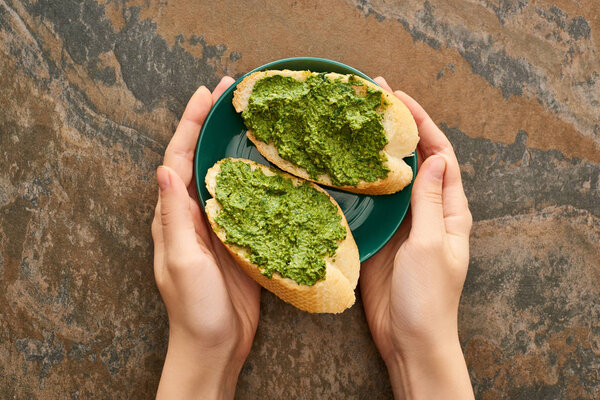 cropped view of woman holding plate with baguette slices and delicious pesto sauce on stone surface