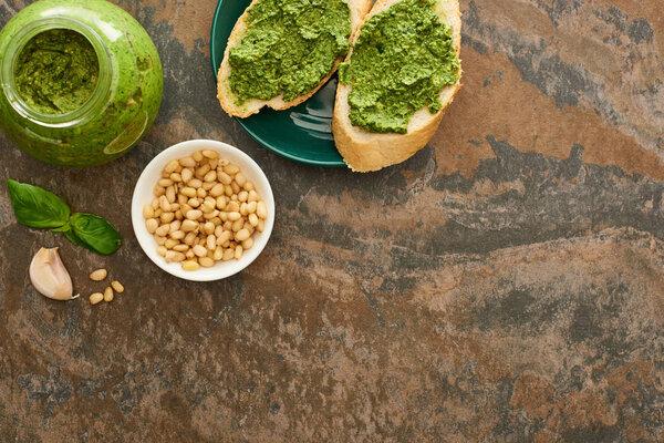 top view of baguette slices with pesto sauce on plate near fresh ingredients on stone surface