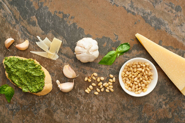 top view of baguette slice with pesto sauce near fresh ingredients on stone surface