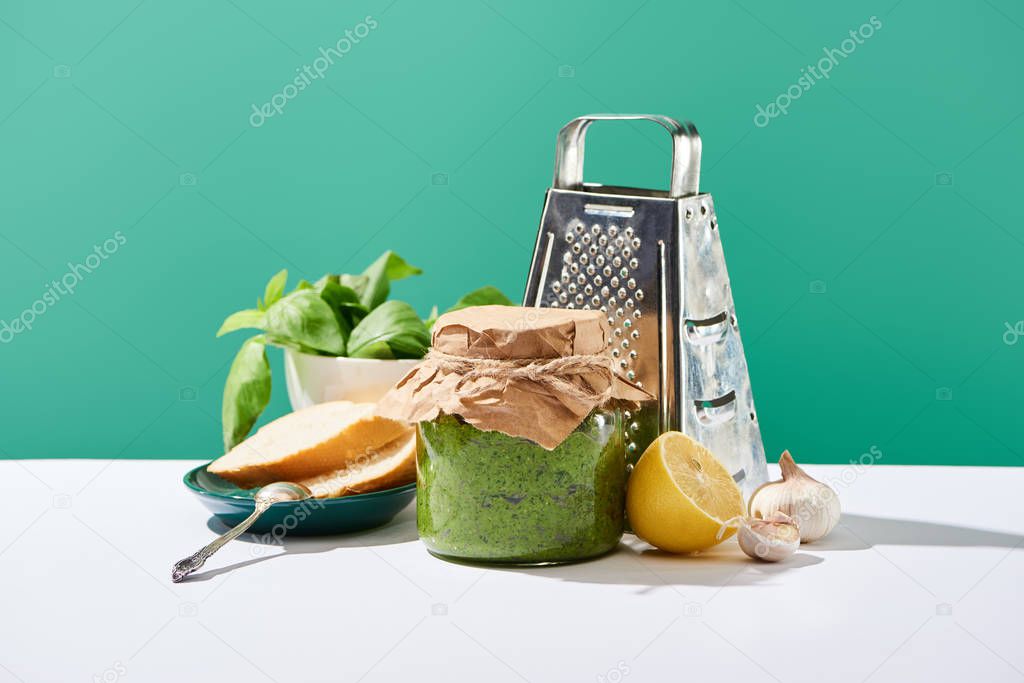 pesto sauce in jar near ingredients, baguette and grater on white table isolated on green