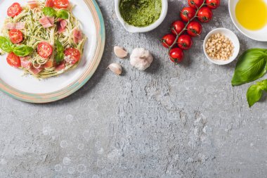 Gri yüzeydeki malzemelerin yanında domates, fesleğen ve prosciutto ile pişmiş Pappardelle 'in üst görüntüsü