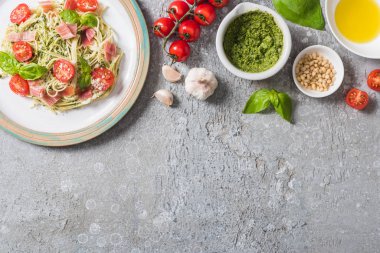 Gri yüzeydeki taze malzemelerin yanında domates, fesleğen ve prosciutto ile pişmiş Pappardelle 'in üst görüntüsü