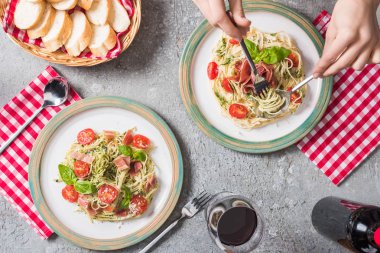 Ekose peçetelerin yanında domatesli, fesleğenli ve jambonlu Pappardelle yiyen bir kadın, gri yüzeyde kırmızı şarap.