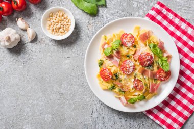 Domates, fesleğen ve prosciutto ile pişmiş Pappardelle 'in gri yüzeydeki malzemelerin yanında ekose peçetenin üzerinde üst görünümü