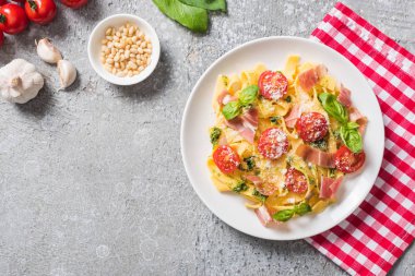 top view of cooked Pappardelle with tomatoes, basil and prosciutto on plaid napkin near ingredients on grey surface clipart