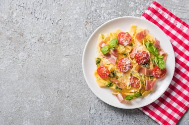 Üzerinde domates, fesleğen ve prosciutto olan pişmiş Pappardelle 'in gri yüzey üzerindeki ekose peçetesinin üst görüntüsü.