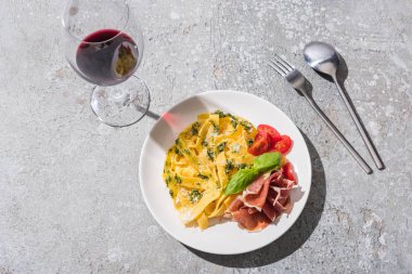 Gri yüzeyde kırmızı şarap ve çatal bıçak yanında domates, pesto ve prosciutto ile lezzetli Pappardelle manzarası