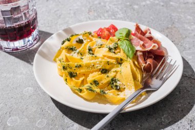 Domatesli lezzetli Pappardelle, pesto ve çatallı prosciutto. Gri yüzeyde kırmızı şarap.
