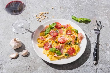 Kırmızı şarap ve gri yüzeydeki malzemeler yanında domates, pesto ve prosciutto ile pappardelle.