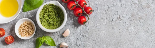 top view of raw tomatoes, garlic, basil, pine nuts, olive oil, pesto sauce on grey surface, panoramic shot