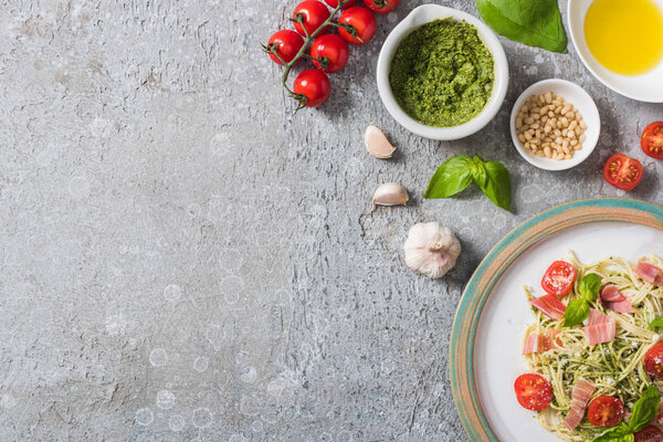top view of cooked Pappardelle with tomatoes, basil and prosciutto near fresh ingredients on grey surface