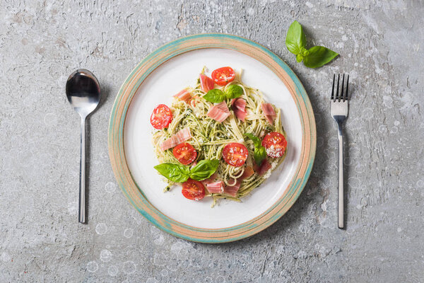 top view of cooked Pappardelle with tomatoes, basil and prosciutto near cutlery on grey surface