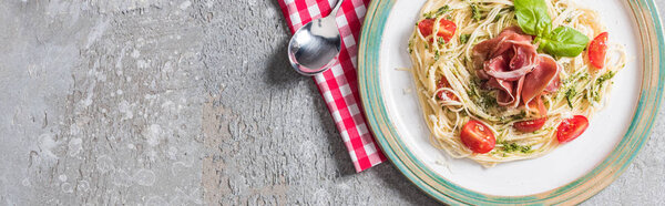 top view of Pappardelle with tomatoes, basil and prosciutto on plate on plaid napkin with spoon on grey surface, panoramic shot