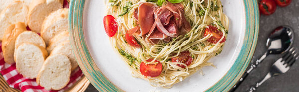 selective focus of served Pappardelle with tomatoes, basil and prosciutto near baguette, tomatoes and cutlery on grey surface, panoramic shot