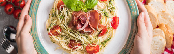 selective focus of woman holding plate with Pappardelle with tomatoes, basil and prosciutto near baguette, tomatoes and cutlery on grey surface, panoramic shot