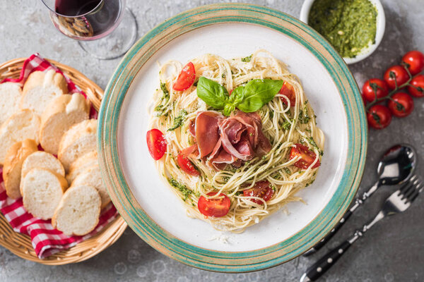 selective focus of served Pappardelle with tomatoes, basil and prosciutto near baguette, red wine, pesto, tomatoes and cutlery on grey surface