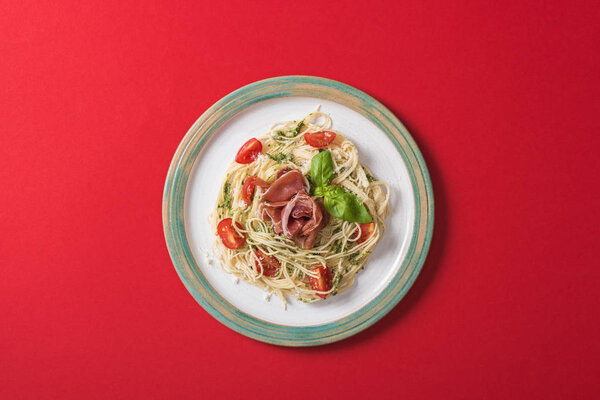 top view of cooked Pappardelle with tomatoes, basil and prosciutto on plate on red background
