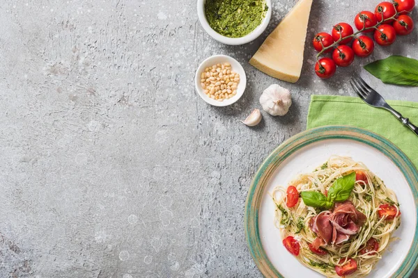 top view of cooked Pappardelle with tomatoes, basil and prosciutto near ingredients on grey surface