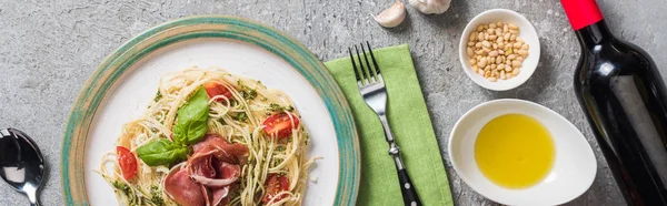 top view of cooked Pappardelle with tomatoes, basil and prosciutto near ingredients, red wine and cutlery on grey surface, panoramic shot