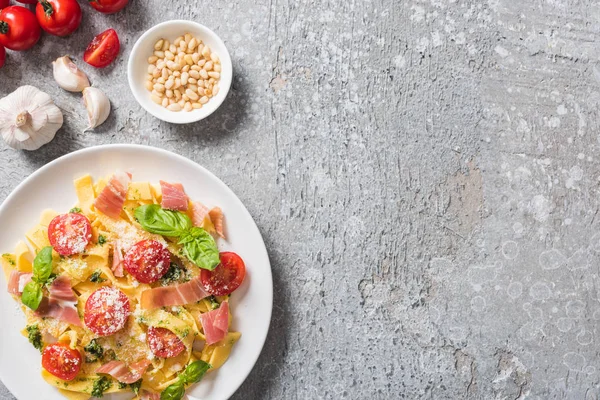 top view of cooked Pappardelle with basil and prosciutto near garlic, pine nuts and tomatoes on grey surface