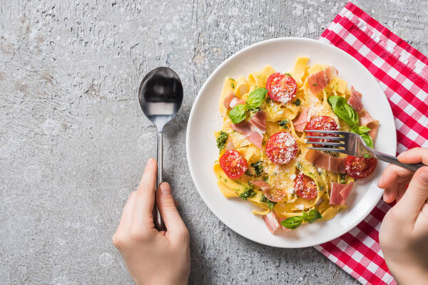 partial view of woman eating Pappardelle with tomatoes, basil and prosciutto on plaid napkin on grey surface
