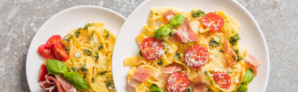 selective focus of cooked Pappardelle with tomatoes, basil and prosciutto on plates on grey surface, panoramic shot