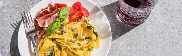top view of tasty Pappardelle with tomatoes, pesto and prosciutto with fork near red wine on grey surface, panoramic shot