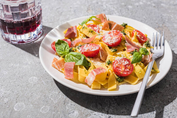 tasty Pappardelle with tomatoes, pesto and prosciutto with fork near red wine on grey surface