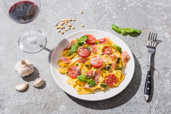 Pappardelle with tomatoes, pesto and prosciutto near red wine and ingredients on grey surface