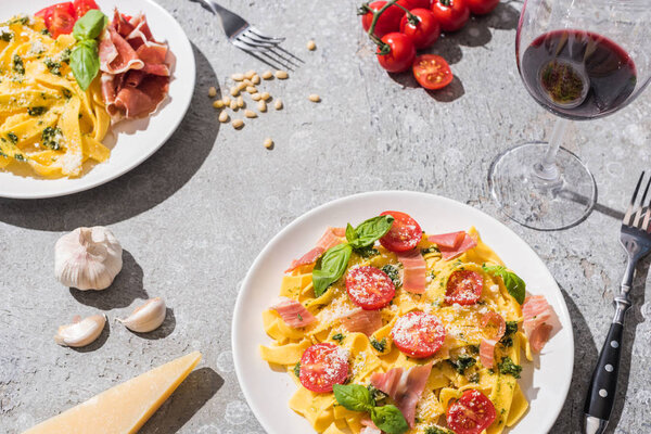 fresh Pappardelle with tomatoes, pesto and prosciutto near red wine and ingredients on grey surface