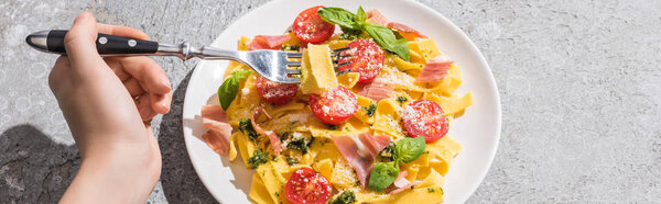 cropped view of woman eating tasty Pappardelle with tomatoes, pesto and prosciutto with fork on grey surface, panoramic shot