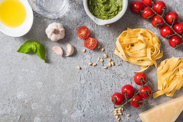 top view of raw Pappardelle near tomatoes, garlic, basil, pine nuts, olive oil, water and pesto sauce on grey surface