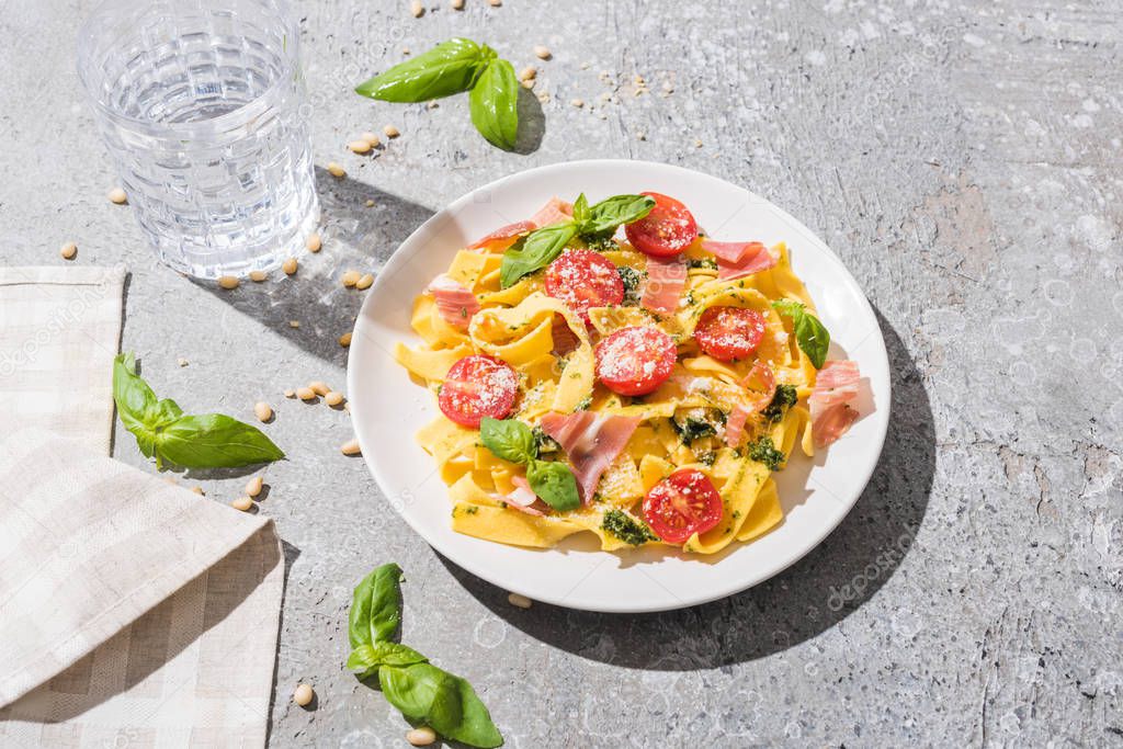 tasty Pappardelle with tomatoes, pesto and prosciutto near water, napkin and pine nuts on grey surface
