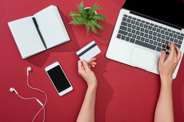 Kredi kartı tutan ve bilgisayarını akıllı telefon, kulaklık, kalem, defter ve kırmızı arka planda bitkilerin yanında kullanan bir kadın görüntüsü.