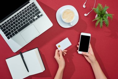 Kahve, dizüstü bilgisayar, kulaklık, not defteri ve kırmızı arka planda bitkinin yanında kredi kartı ve akıllı telefon tutan kadının kısmi görüntüsü