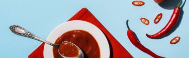 Top view of delicious spicy ketchup in plate with ripe chili peppers on blue surface, panoramic shot clipart