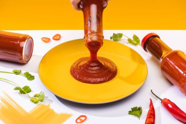 Cropped view of woman pouring ketchup on plate beside spaghetti and chili peppers on white surface isolated on yellow clipart