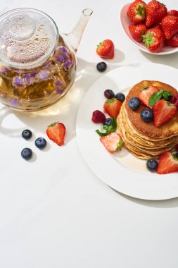 top view of delicious pancakes with maple syrup, blueberries and strawberries on plate near herbal tea in teapot on marble white surface clipart
