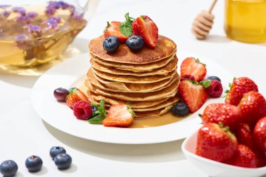 selective focus of delicious pancakes with honey, blueberries and strawberries on plate on white surface clipart
