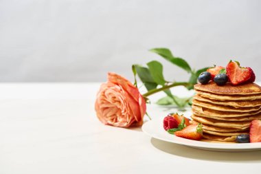 selective focus of delicious pancakes with blueberries and strawberries on plate near rose on white surface isolated on grey clipart