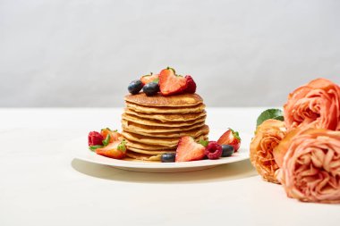 selective focus of delicious pancakes with blueberries and strawberries on plate near rose flowers on white surface isolated on grey