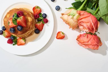 top view of delicious pancakes with honey, blueberries and strawberries on plate near roses on marble white surface clipart