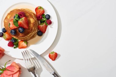 top view of delicious pancakes with honey, blueberries and strawberries on plate near cutlery and rose on marble white surface clipart