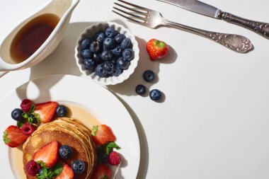 top view of delicious pancakes with blueberries and strawberries on plate near cutlery and maple syrup in gravy boat on marble white surface clipart