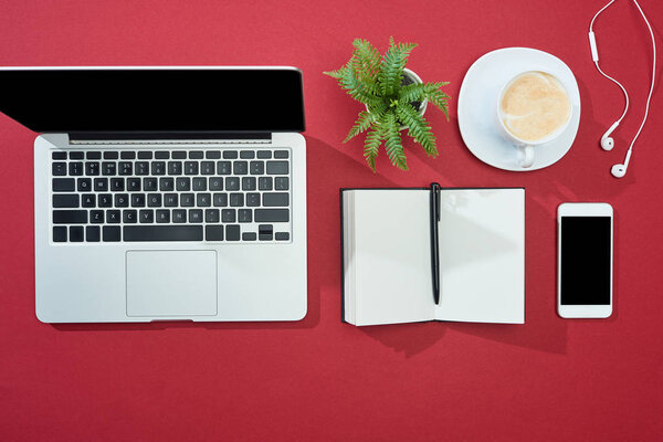 flat lay with smartphone, laptop, earphones, coffee, notebook with pen and plant on red background