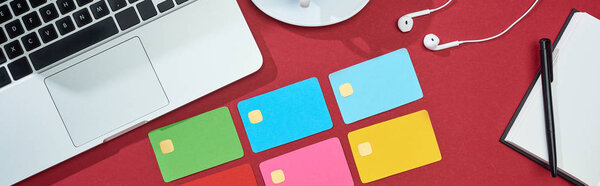 вид сверху на многоцветные пустые кредитные карты на красном фоне с ноутбуком, наушниками и ноутбуком, панорамный снимок
