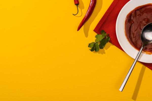 Top view of spicy chili sauce in plate with cilantro and chili peppers on yellow background