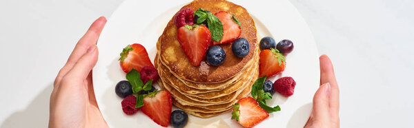 cropped view of woman holding plate with delicious pancakes with honey, blueberries and strawberries, panoramic shot