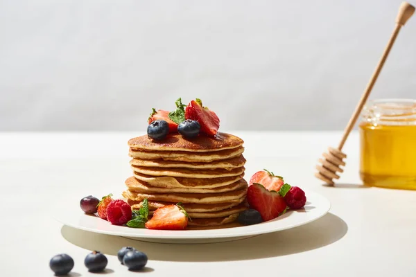 ブルーベリーとイチゴをプレートに盛り付けた美味しいパンケーキをグレーに孤立させた白い表面にハチミツの近くで — ストック写真